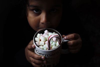 Close-up portrait of boy holding marshmallows in cup against black background