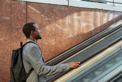 Side view of african american male entrepreneur with backpack listening to songs in tws earbuds on escalator