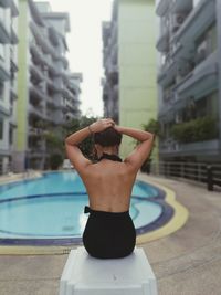 Rear view of young woman tying hair while sitting on stool at poolside