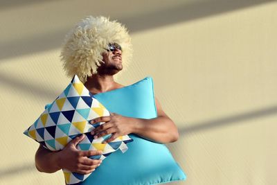 Man wearing wig holding cushions against wall