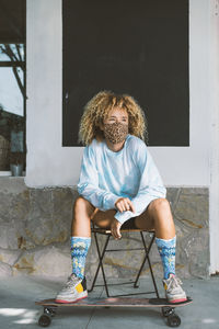 Woman wearing face mask sitting on chair with skateboard against wall