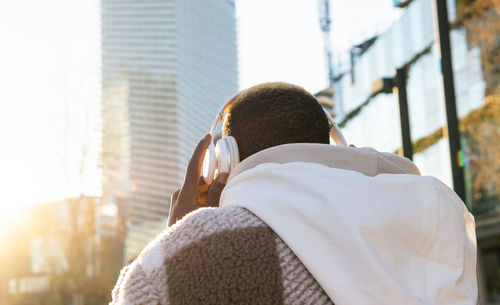 Back view of unrecognizable male with short hair putting on headphones while listening to music on sunny street with modern buildings