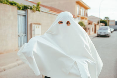 Unrecognizable kid in white ghost costume having fun on street during halloween holiday and looking at camera