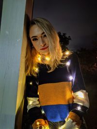 Portrait of young woman standing against illuminated wall at night