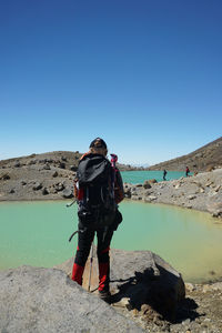 Rear view of woman standing on rock by lake against clear blue sky