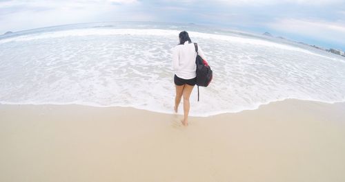 Rear view of woman with backpack walking at beach against sky