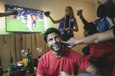 Cropped hand of man applying face paint on friend's cheek during soccer match