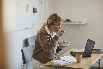 Female professor eating lunch while sitting at desk with laptop in classroom