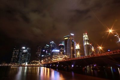 Illuminated modern buildings by river against sky at night