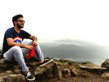 Young man in sunglasses sitting on mountain against sky