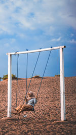 Boy sitting on swing at playground against sky