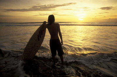 Rear view of shirtless surfer with surfboard at beach against sky during sunset