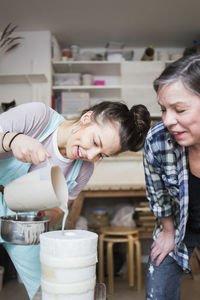 Mature female potter bending while looking at smiling young employee pouring clay from pitcher in vase at workshop