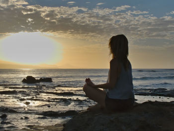 Woman sitting on shore at beach against sky during sunset