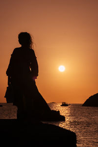 Rear view of silhouette woman standing at beach against sky during sunset