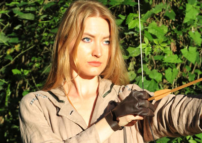 Close-up of confident young woman aiming bow in forest during sunny day