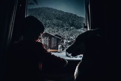 Rear view of boy sitting with dog by window at home