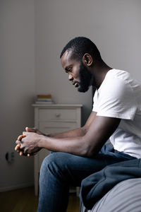 Side view of depressed lonely man sitting with hands clasped on bed at home