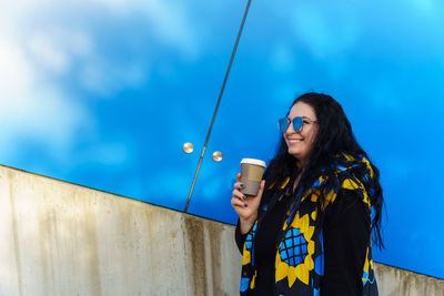 Woman with long curly hair in black, blue and yellow waistcoat drinks coffee from paper cup