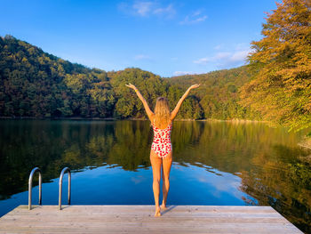 Rear view of young woman in swimming suit standing on a wooden pontoon near a lake 