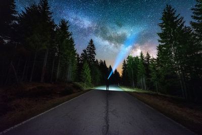 Silhouette man standing on road amidst trees in forest against star field at night