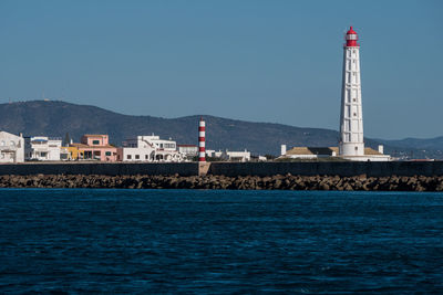 Lighthouse by sea and buildings against clear sky