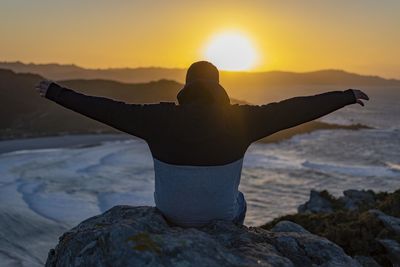 Rear view of man with arms outstretched sitting on rock at beach during sunset