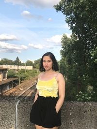 Portrait of young woman standing by retaining wall against sky