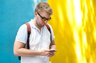 Young man looking at phone while standing against blue yellow wall