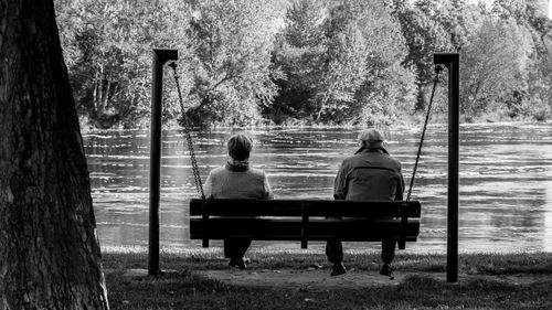 Rear view of old man and woman sitting on wooden swing by lake