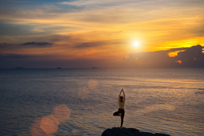 Rear view of woman meditating while standing by sea against sky during sunset