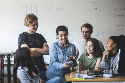 Surprised teacher examining robot while sitting amidst students against whiteboard in classroom at high school