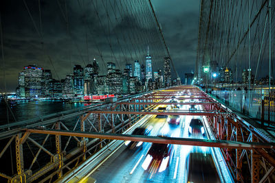 Light trails on bridge by buildings against sky at night