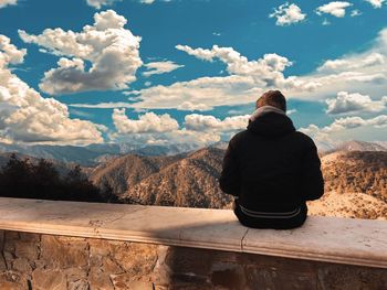 Rear view of man sitting on retaining wall against mountain ranges and cloudy sky