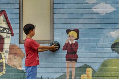 Boy holding ball while standing by mural wall
