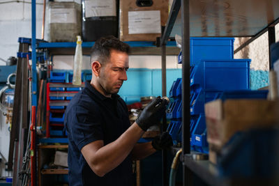 Positive adult male master in protective gloves picking metal detail from box while choosing tools for repair service in workshop