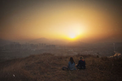 Rear view of friends siting on mountain peak by city against sky during sunset