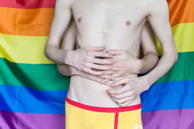 Midsection of gay couple embracing while standing against rainbow flag