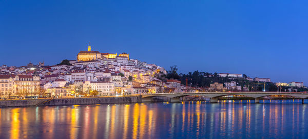 Illuminated buildings by river against clear blue sky at dusk