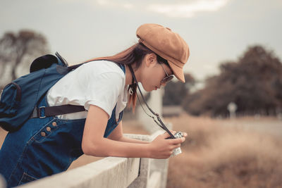 Side view of young woman photographing with camera while leaning on railing