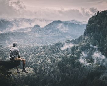 Rear view of hiker sitting on cliff against sky