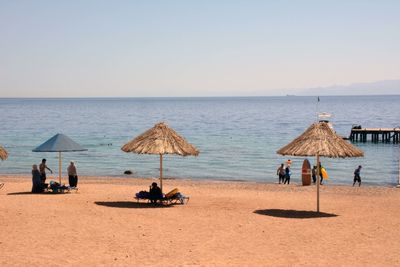 People and thatched roofs at beach against clear sky