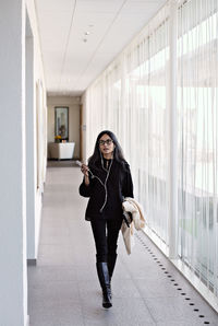 Full length businesswoman talking on smart phone while walking in corridor at office