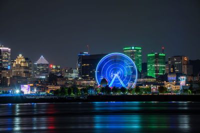 Illuminated ferris wheel by river against buildings at night