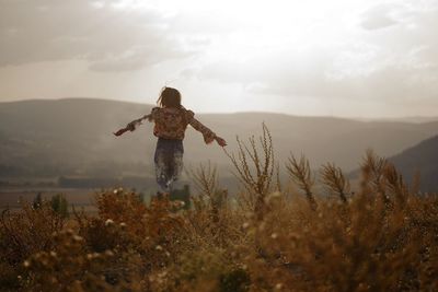 Rear view of woman with arms outstretched levitating over field against sky during sunset