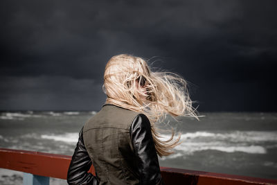 Woman with tousled hair standing by railing at beach against storm clouds