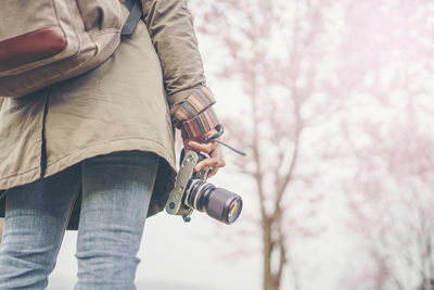 Midsection of woman holding camera while standing against tree