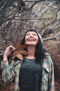 Smiling woman holding leaf while looking up standing against bare trees