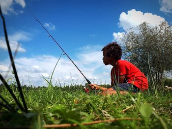 Side view of boy with fishing rod sitting on grassy field against sky