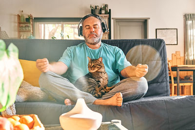 Middle-aged overweight man in wireless headphones relaxing at home with cat and guided meditation.